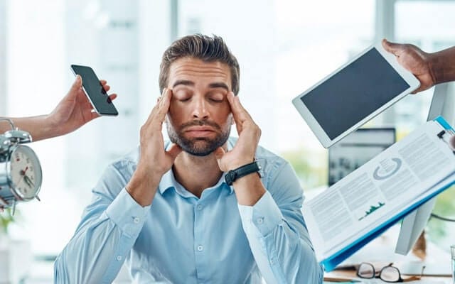 Hombre con los ojos cerrados rodeado de teléfono, tableta, despertador y documentos