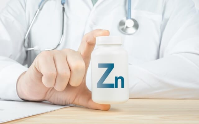 Médico con bata blanca sostiene un frasco de suplemento marcado con el símbolo Zn